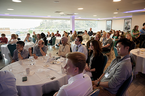 Members of the audience in a conference room, seated around round tables and watching a presentation
